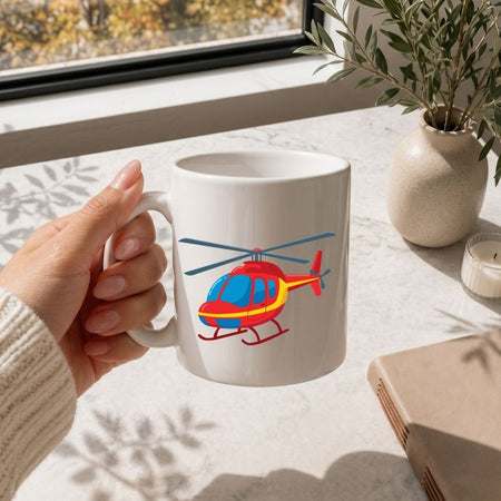 A hand holding a white ceramic mug with a colorful cartoon helicopter graphic, set on a light marble tabletop near a window with a potted plant and a candle in the background.