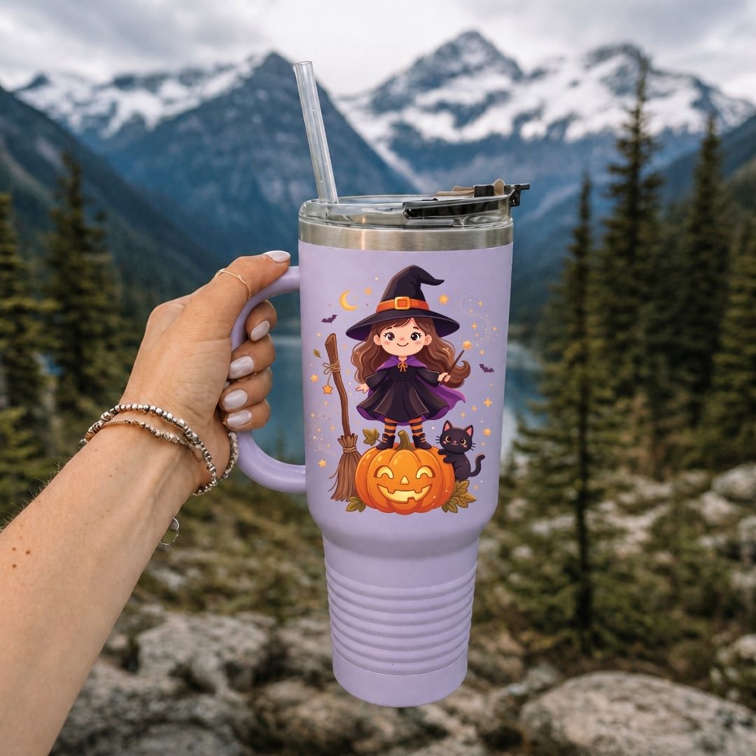 A lavender insulated tumbler with a cartoon witch graphic, held by a hand against a mountain and forest background.
