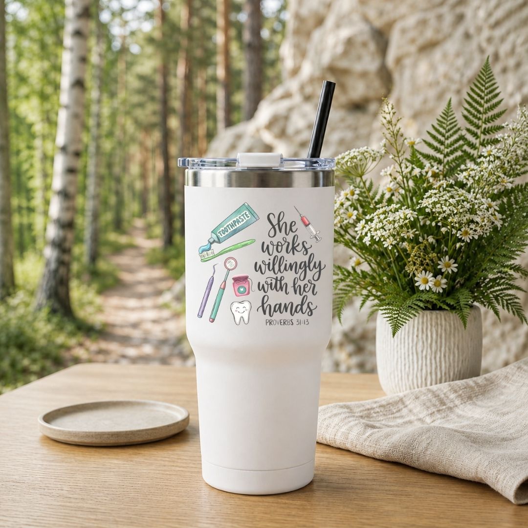 A white insulated tumbler with a clear lid and black straw sits on a wooden table outdoors. The tumbler has a printed message reading 'She works willingly with her hands PHILIPPIANS 9:11' with small dental-themed illustrations.