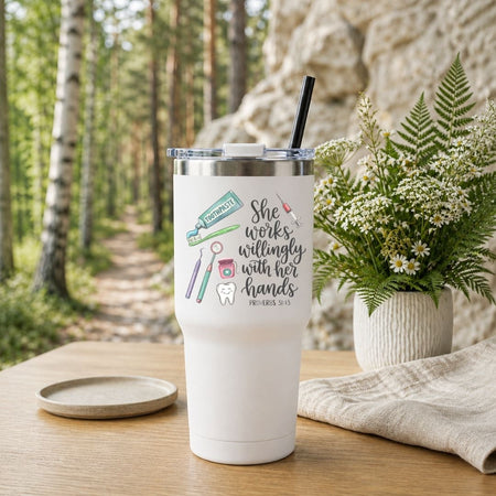 A white insulated tumbler with a clear lid and black straw sits on a wooden table outdoors. The tumbler has a printed message reading 'She works willingly with her hands PHILIPPIANS 9:11' with small dental-themed illustrations.