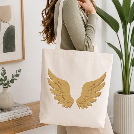 A woman wearing a sage green ribbed sweater carries a cream canvas tote bag with a large gold wing graphic on the front, set in a bright, modern room with a wooden table, books, and a potted plant.