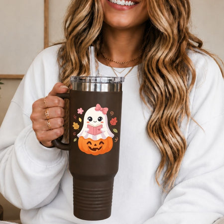 A woman in a white sweatshirt holds a dark brown insulated tumbler with a handle and clear lid and straw. The tumbler has a Halloween graphic featuring a white ghost reading a pink book while sitting on an orange jack-o'-lantern, with small autumn leaves around it.