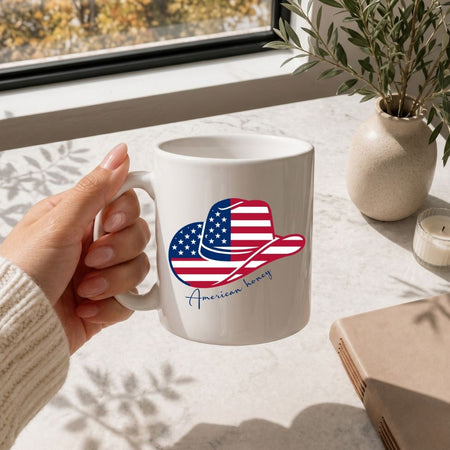 A hand with polished nails holding a clean white ceramic coffee cup decorated with the American flag cowboy hat design on a marble table.
