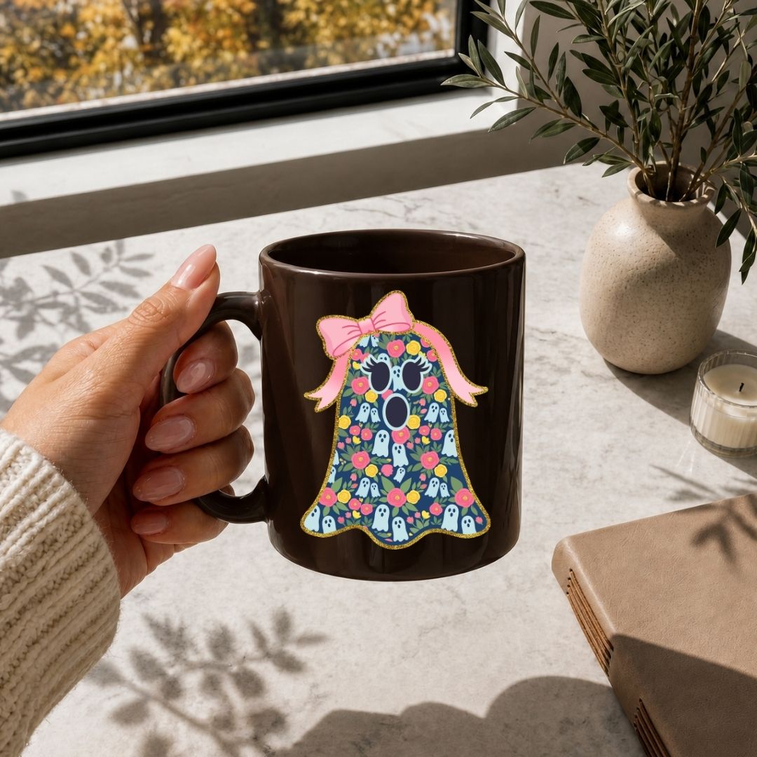 A hand holds a dark brown ceramic mug featuring a colorful graphic of a ghost with a pink bow and floral pattern. The scene is set on a light stone tabletop near a window with a plant and candle in the background.