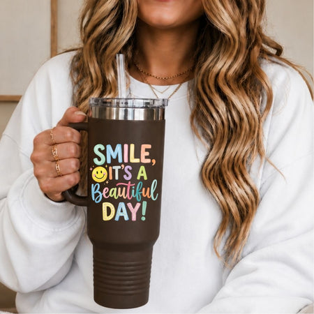 A woman in a white sweatshirt holds a dark brown insulated tumbler with a handle, clear lid, and straw. The tumbler is printed with the phrase “SMILE, IT’S A Beautiful DAY!” in colorful lettering with a yellow smiley face.