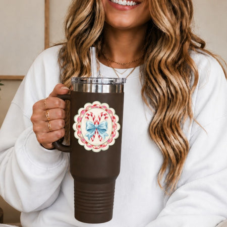 A woman in a white sweatshirt holds a dark brown insulated tumbler with a clear straw. The tumbler has a silver lid and a decorative circular holiday design featuring candy canes and a blue bow.