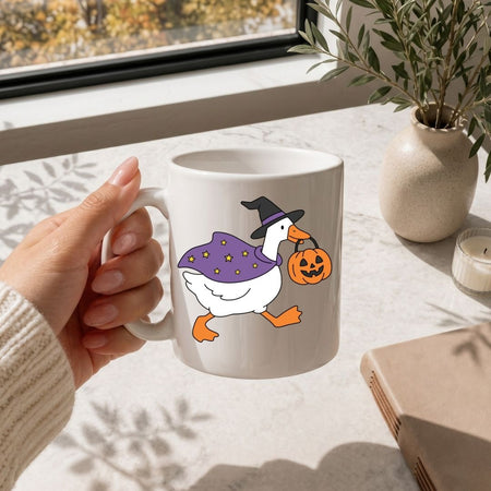 A hand holding a white ceramic mug with a cartoon duck wearing a purple witch hat and cape while carrying a jack-o'-lantern, set in a bright room near a window with a plant and candle in the background.