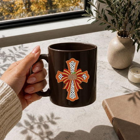 A hand holding a dark brown ceramic mug with a decorative orange, red, and white cross design, set in a sunlit room near a window with a plant and candle in the background.