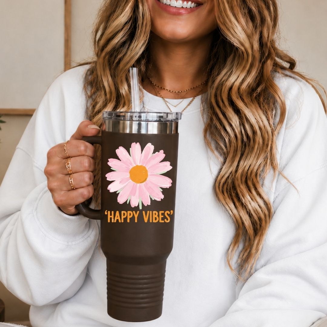 A woman in a white sweatshirt holds a dark brown insulated tumbler with a pink daisy graphic and the words “HAPPY VIBES” in yellow. The tumbler has a clear lid with a metal straw.