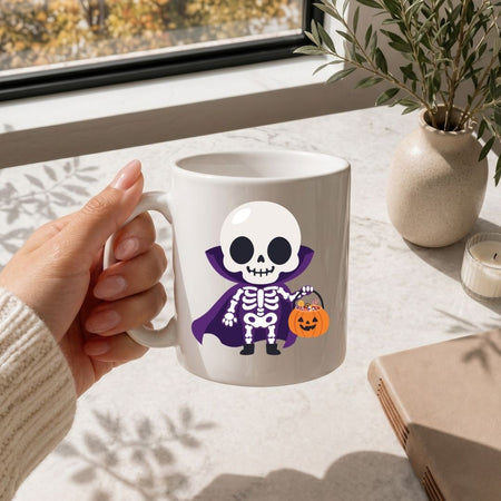 A hand holding a white ceramic mug with a cartoon skeleton wearing a purple cape and holding an orange pumpkin bucket, set on a light marble tabletop near a window with a plant and candle in the background.