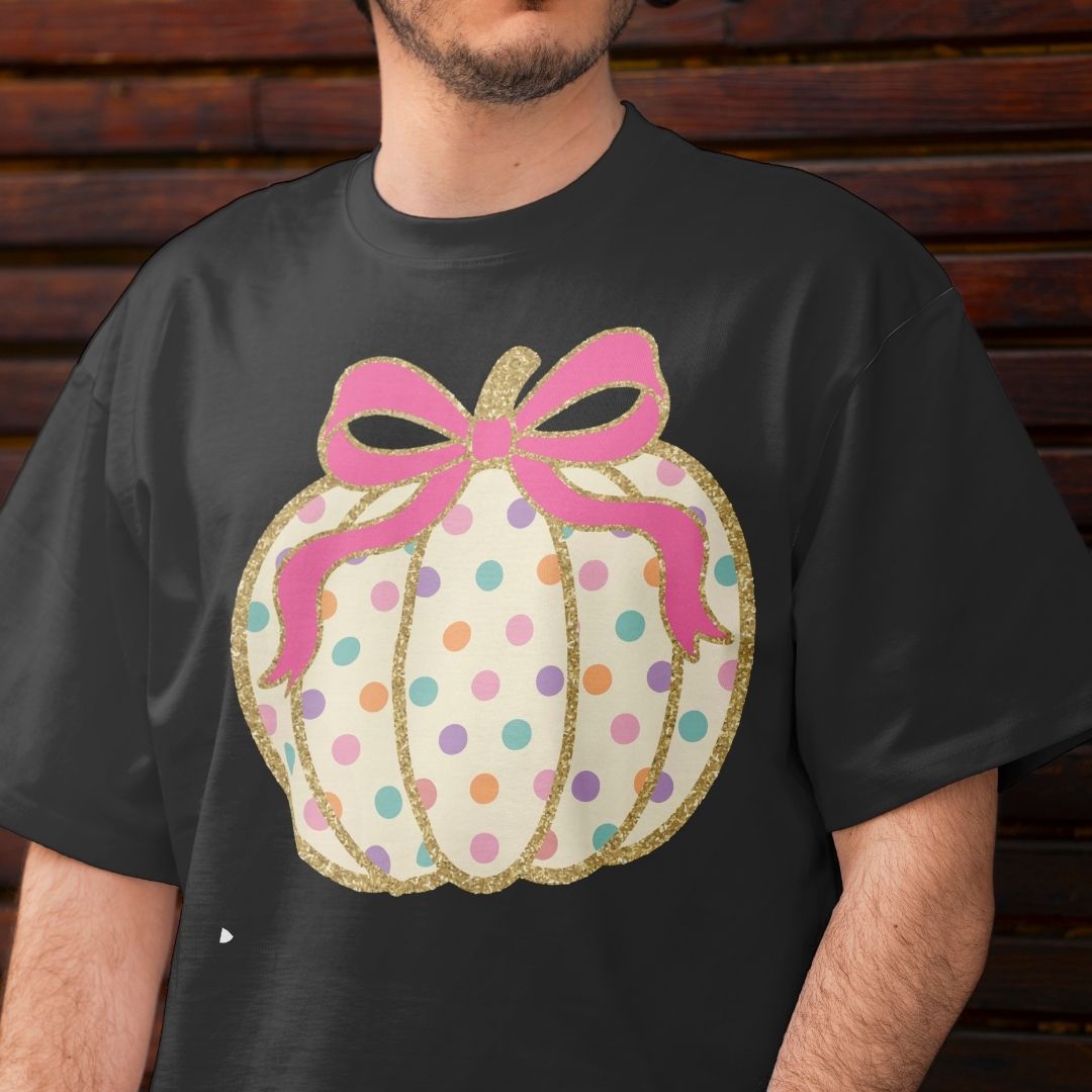 A man wearing a black t-shirt with a graphic of a pumpkin-shaped gift box decorated with pink glittery ribbon, gold glitter trim, and pastel-colored polka dots on a cream background.