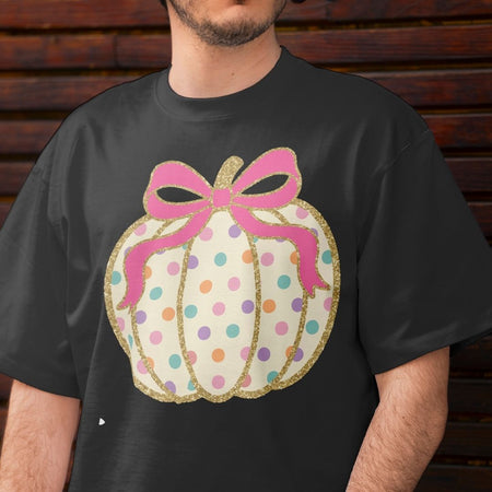 A man wearing a black t-shirt with a graphic of a pumpkin-shaped gift box decorated with pink glittery ribbon, gold glitter trim, and pastel-colored polka dots on a cream background.