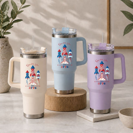 Three insulated travel tumblers in cream, light blue, and lavender purple arranged on a table, all showcasing the "Sparkle Like It's 1776" rocket design.