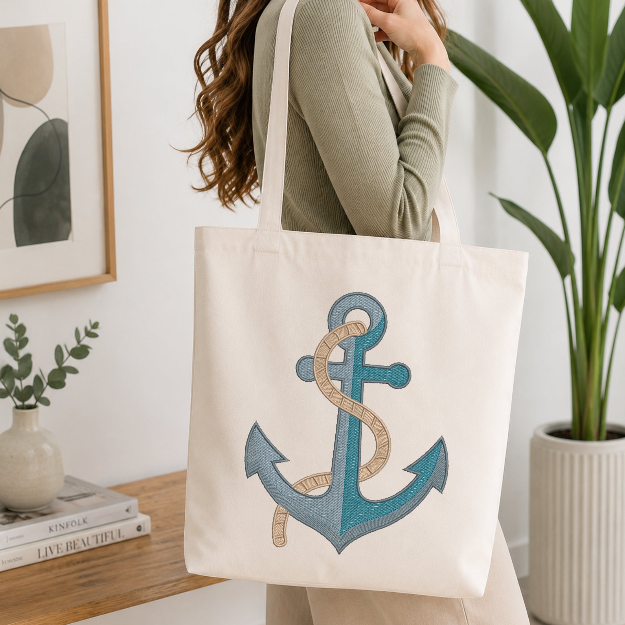 A woman wearing a sage green ribbed sweater carries a cream canvas tote bag with a large embroidered anchor design in shades of blue and beige.