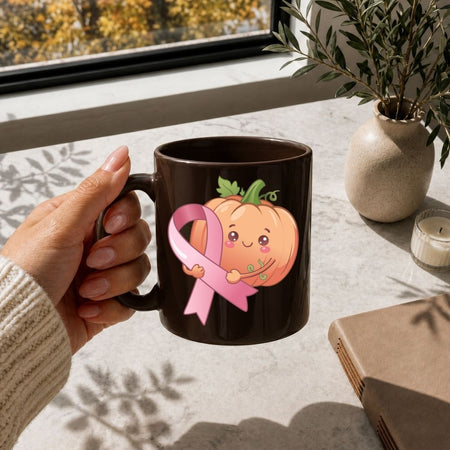A hand holds a dark brown ceramic mug featuring a cute cartoon pumpkin and pink ribbon graphic, set in a sunlit room with a window, potted plant, candle, and book nearby.