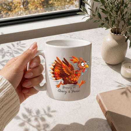 A hand holding a white ceramic mug with a colorful cartoon turkey graphic and the text 'Running on Gray & Panic' on a sunlit marble countertop near a window and a small vase.