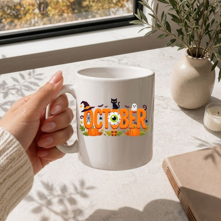 Hand holding a white coffee mug printed with a festive "October" Halloween design featuring pumpkins and a witch hat.