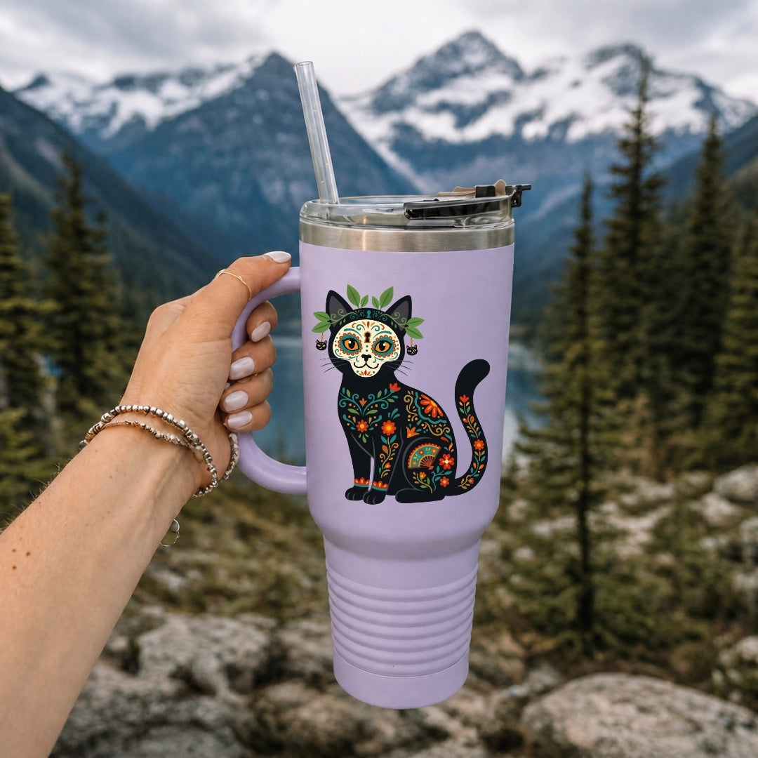 A hand holds a lavender insulated tumbler with a clear straw in front of a mountain and forest background. The tumbler features a colorful sugar-skull cat graphic with floral patterns.