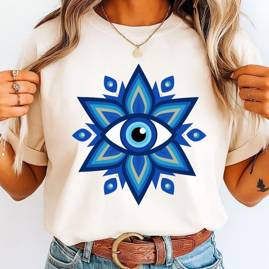 A woman wearing a cream-colored t-shirt with a large blue and teal geometric evil eye design on the front, paired with blue jeans and a brown leather belt.
