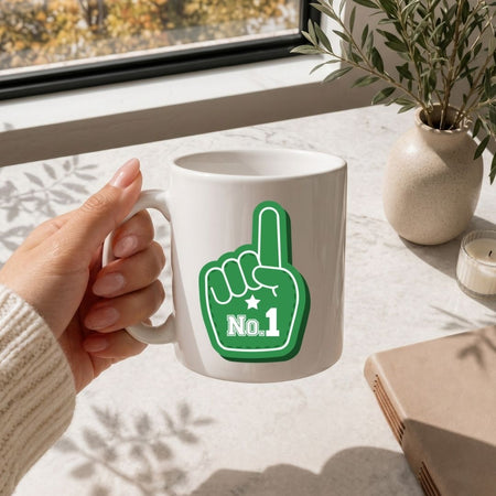 A hand holding a white ceramic mug with a green foam finger graphic that says 'No.1' in white, set on a light marble tabletop near a window with a potted plant and candle in the background.