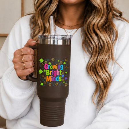 A woman wearing a white sweatshirt holding a dark brown insulated travel tumbler cup printed with a colorful Growing Bright Minds graphic design featuring stars and smiley faces.