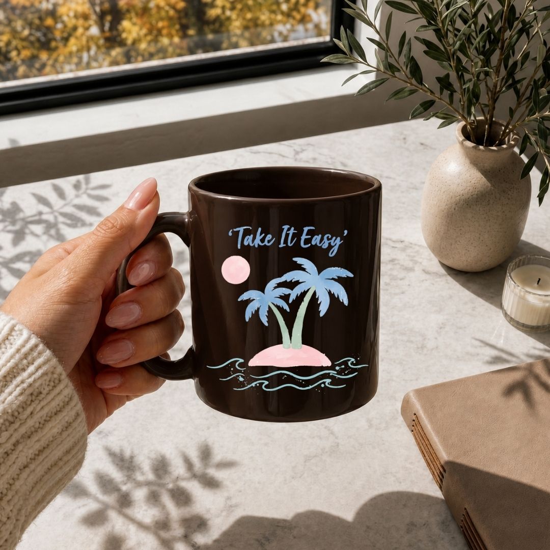 A hand holds a dark brown ceramic mug with a tropical island graphic and the text 'Take It Easy' in a bright lifestyle setting near a window.