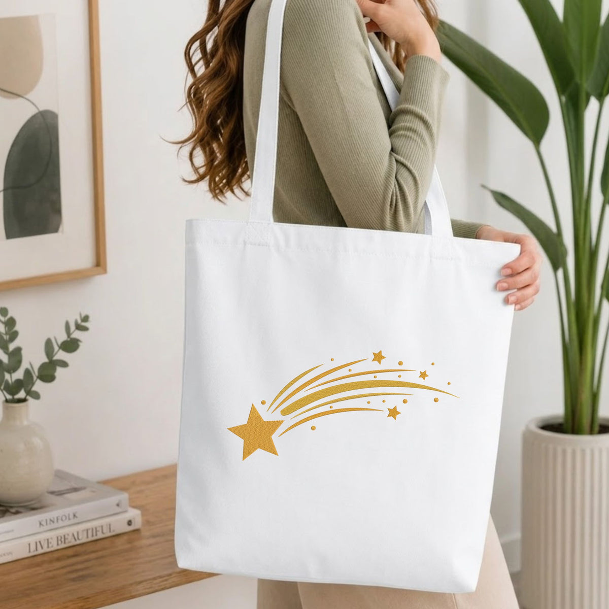 A white canvas tote bag with a gold shooting star graphic, carried by a woman wearing a sage green ribbed sweater in a bright, modern room with a potted plant and books on a wooden table.