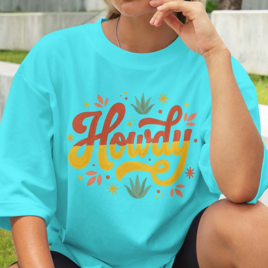 Person wearing a turquoise t-shirt with 'Howdy' print in red and yellow, sitting outdoors.