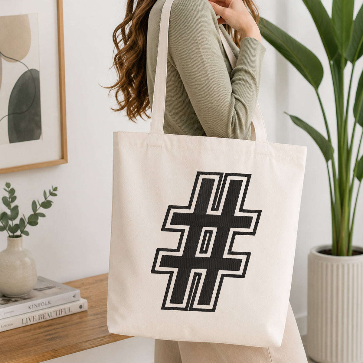 A woman wearing a sage green ribbed long-sleeve top carries a large off-white canvas tote bag with a bold black geometric hashtag symbol printed on the front, set in a bright, modern room with a wooden table, framed art, and a potted plant.