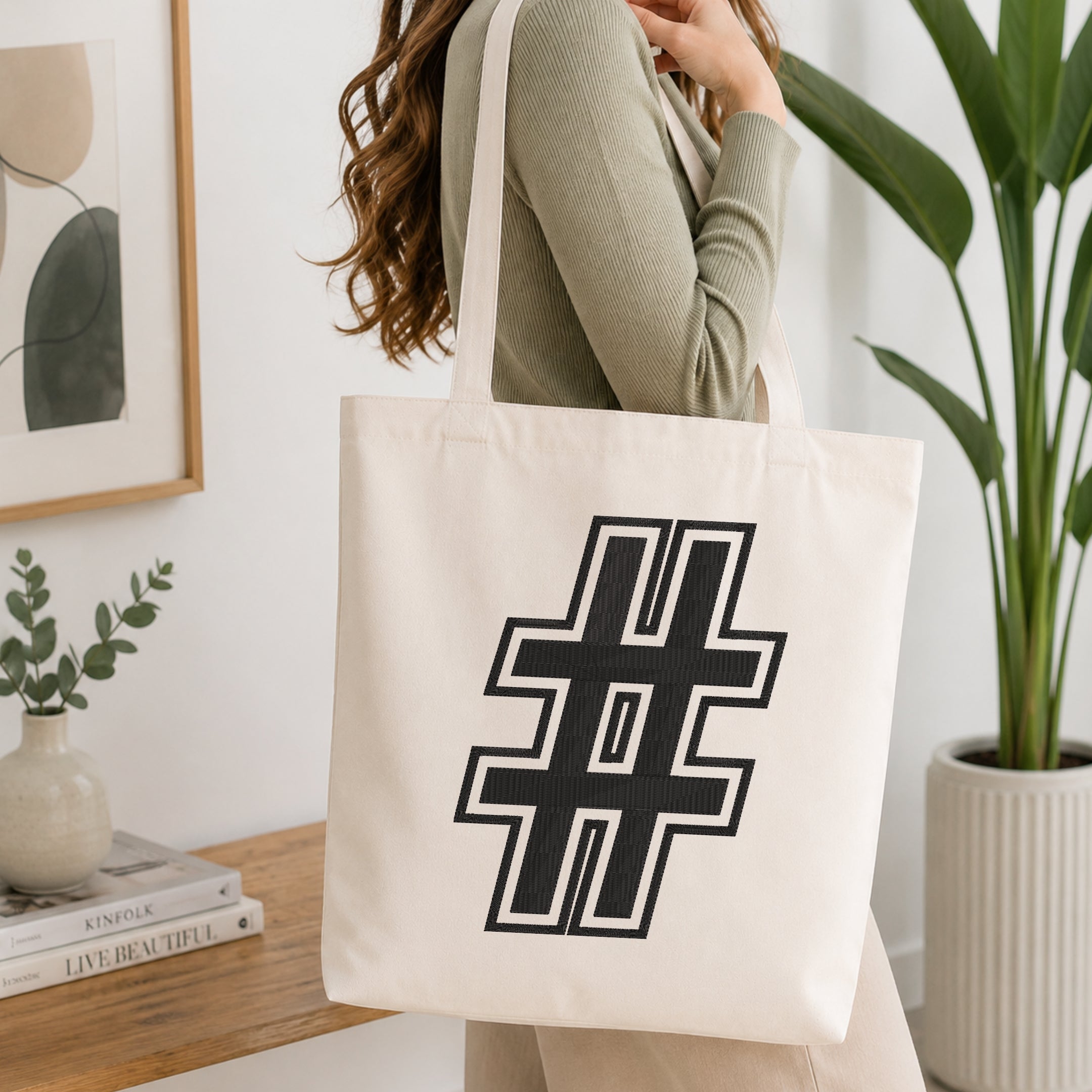 A woman wearing a sage green ribbed long-sleeve top carries a large off-white canvas tote bag with a bold black geometric hashtag symbol printed on the front, set in a bright, modern room with a wooden table, framed art, and a potted plant.