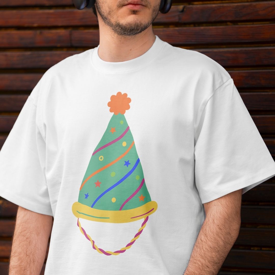 A man wearing an off-white t-shirt printed with a green party hat decorated with colorful wavy lines, stars, dots, and an orange pom-pom.