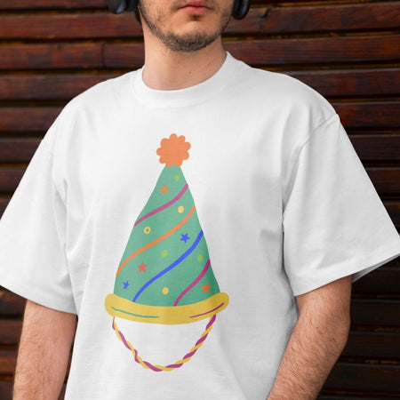 A man wearing an off-white t-shirt printed with a green party hat decorated with colorful wavy lines, stars, dots, and an orange pom-pom.