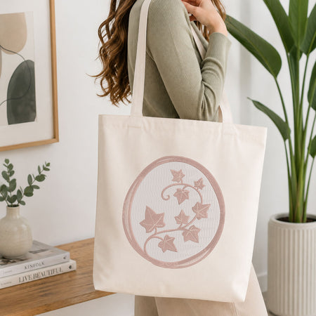 Woman carrying a white natural canvas tote bag featuring an embroidered ivy vine design inside an oval frame in a bright, modern interior setting