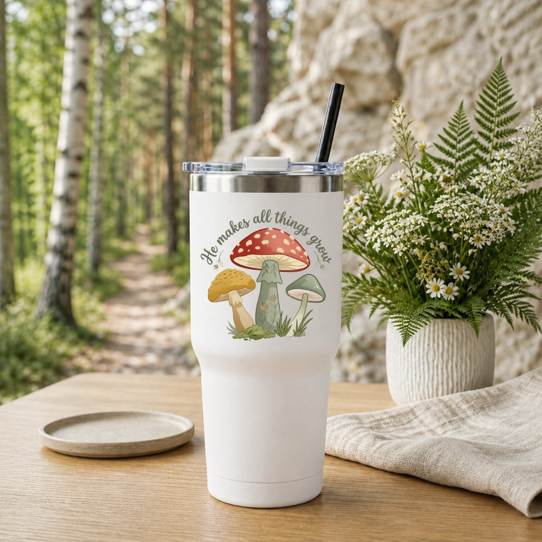 A white insulated tumbler with a clear lid and black straw sits on a wooden table outdoors. The tumbler has a printed mushroom illustration and the phrase “He makes all things grow.