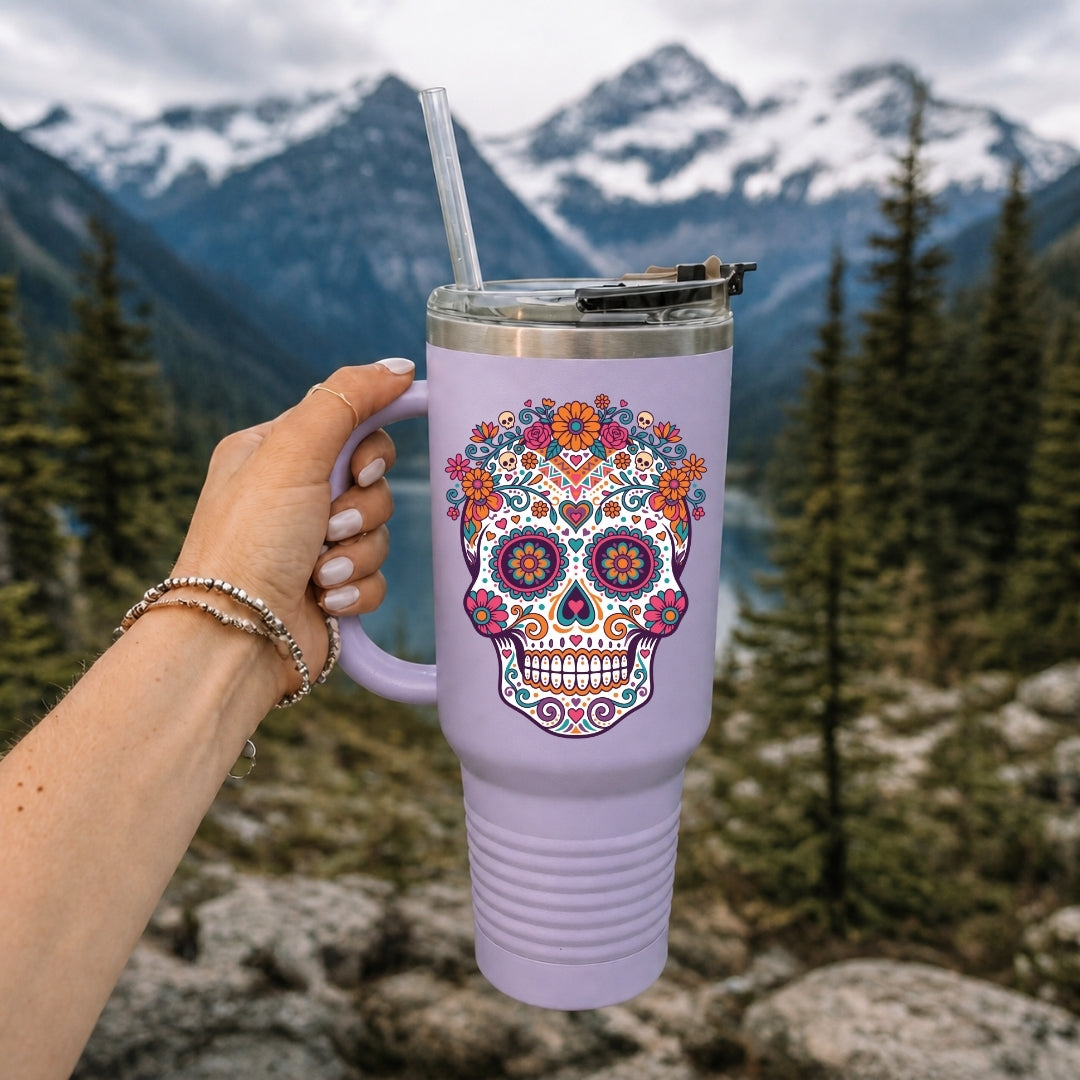 A lavender insulated tumbler with a colorful sugar skull design, held by a hand against a mountain and forest background.