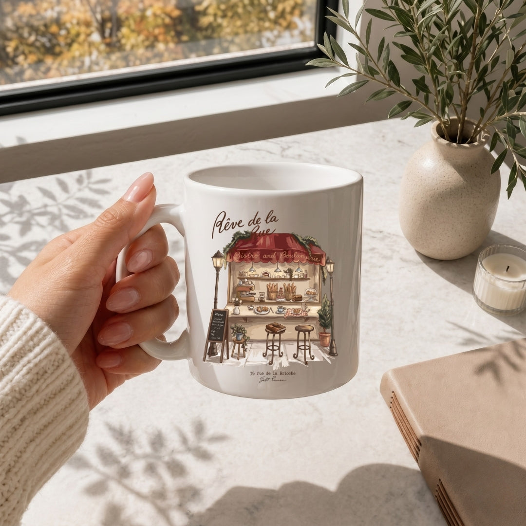 Hand holding a white ceramic coffee mug printed with the charming French bakery stall illustration.