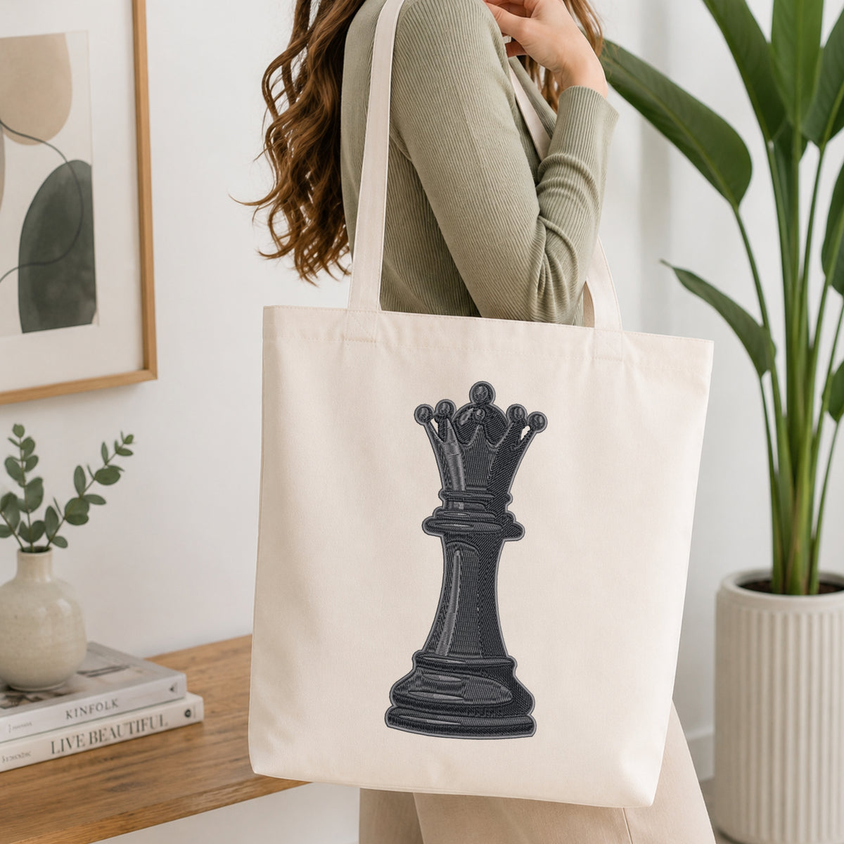 A woman wearing a sage green ribbed sweater carries a cream canvas tote bag with a large black chess queen graphic on the front, set in a bright, modern room with a wooden table, framed art, and a potted plant.