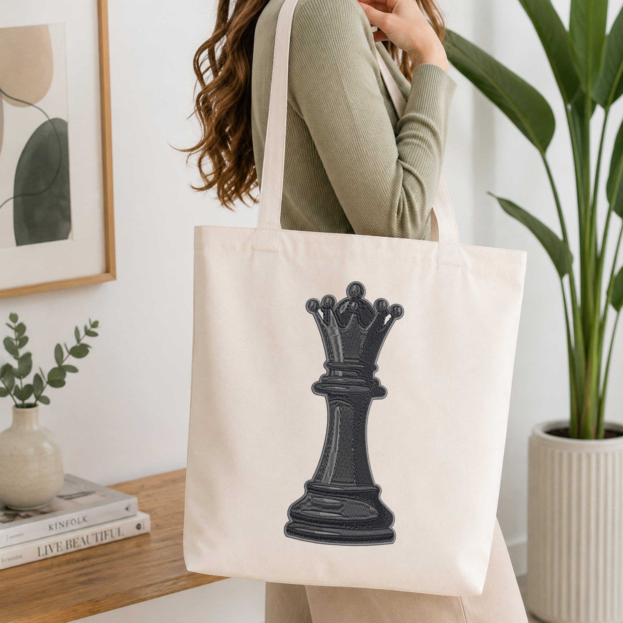 A woman wearing a sage green ribbed sweater carries a cream canvas tote bag with a large black chess queen graphic on the front, set in a bright, modern room with a wooden table, framed art, and a potted plant.