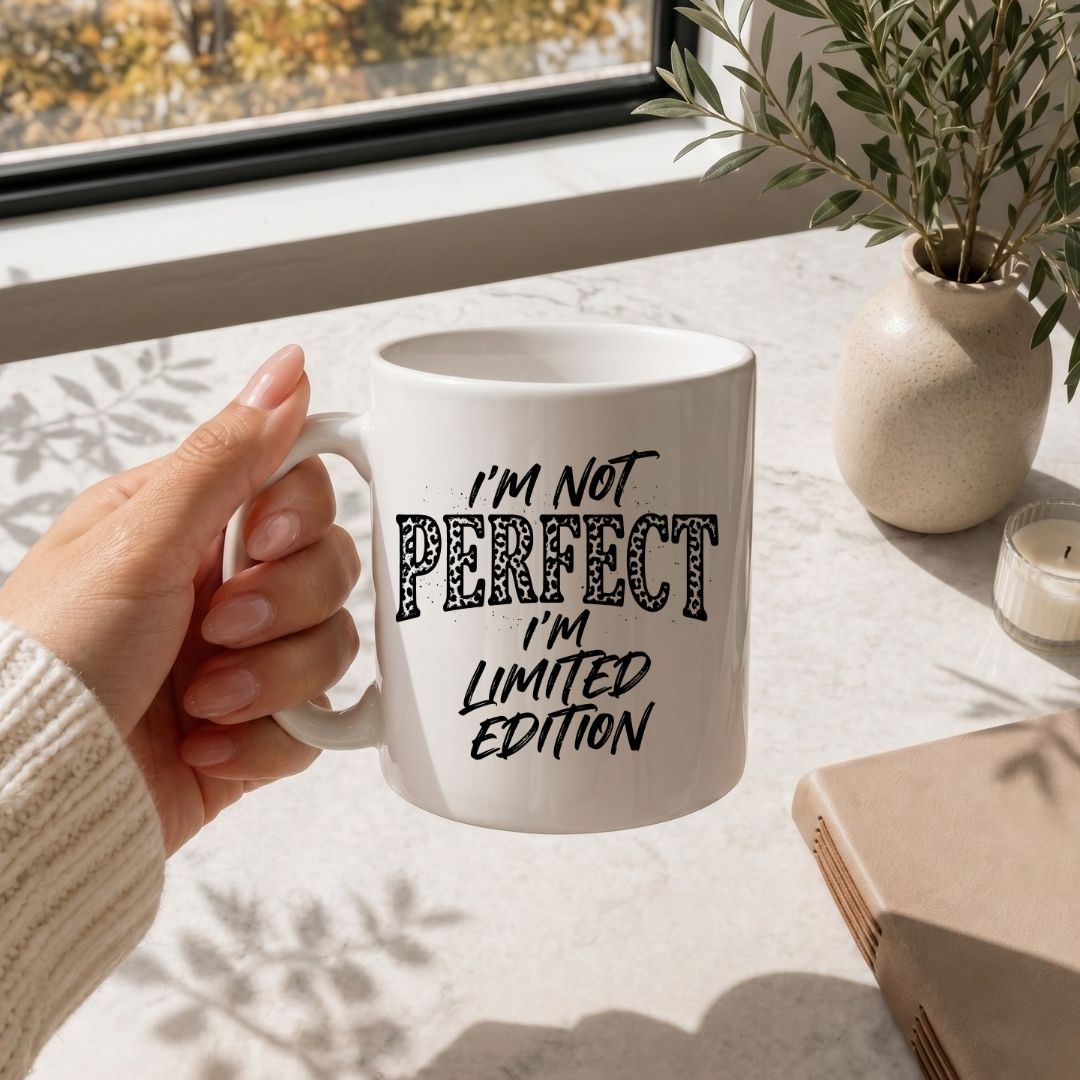 A hand holding a white ceramic mug with the text 'I'M NOT PERFECT I'M LIMITED EDITION' in black lettering, set on a marble tabletop near a window with a plant and candle in the background.
