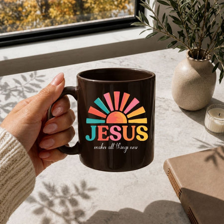 A hand holds a dark brown ceramic mug with a colorful sunburst graphic and the text 'JESUS makes all things new' in front of a window, with a potted plant and candle in the background.