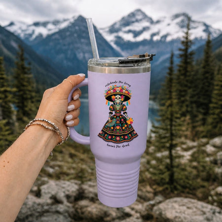 A hand holds a lavender insulated tumbler with a clear straw in front of a blurred mountain and forest background. The tumbler has a colorful illustrated graphic of a decorated skeleton figure and small text.