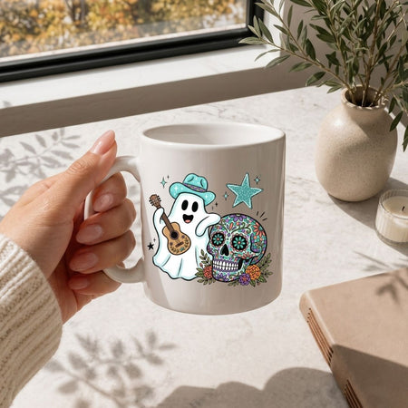 A person holding a white ceramic coffee mug printed with the ghost and sugar skull Halloween illustration, placed on a marble surface near a vase of greenery.