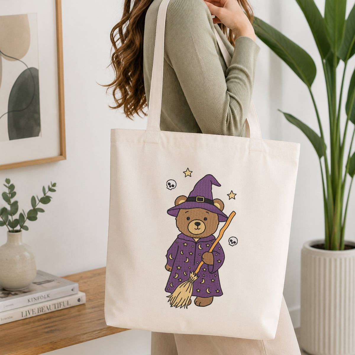 A woman wearing a sage green ribbed sweater carries a cream canvas tote bag. The bag features a cute cartoon illustration of a teddy bear dressed as a witch in a purple robe and hat, holding a broomstick with small stars and skull-and-crossbones details.