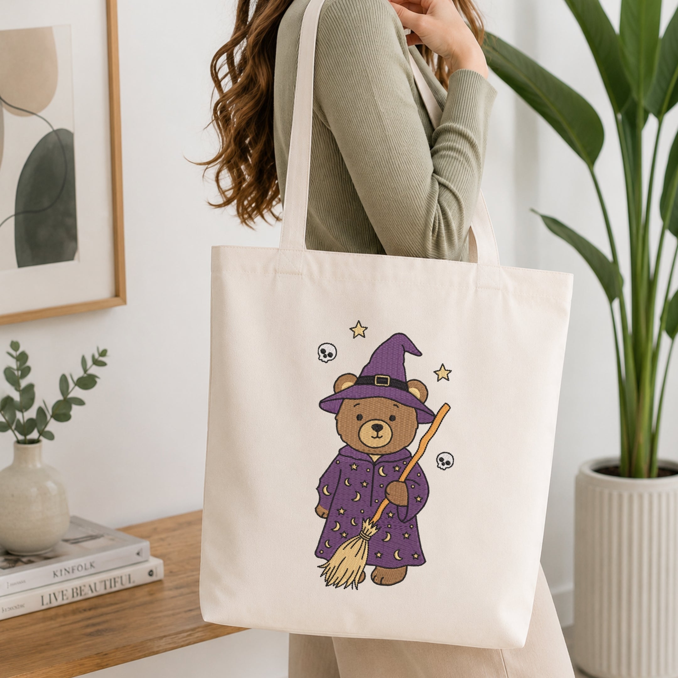 A woman wearing a sage green ribbed sweater carries a cream canvas tote bag. The bag features a cute cartoon illustration of a teddy bear dressed as a witch in a purple robe and hat, holding a broomstick with small stars and skull-and-crossbones details.