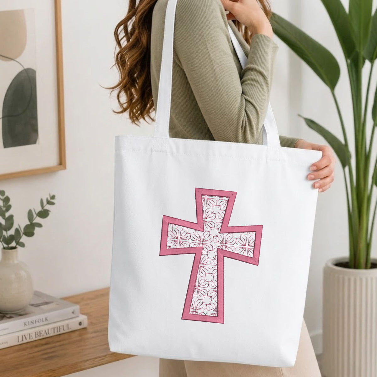 A woman wearing a sage green ribbed sweater carries a white canvas tote bag with a large pink cross design featuring a white floral pattern.
