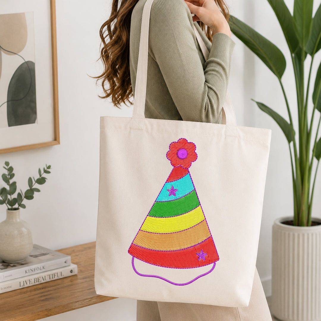 Person holding a canvas tote bag featuring a colorful rainbow striped birthday hat embroidery design.