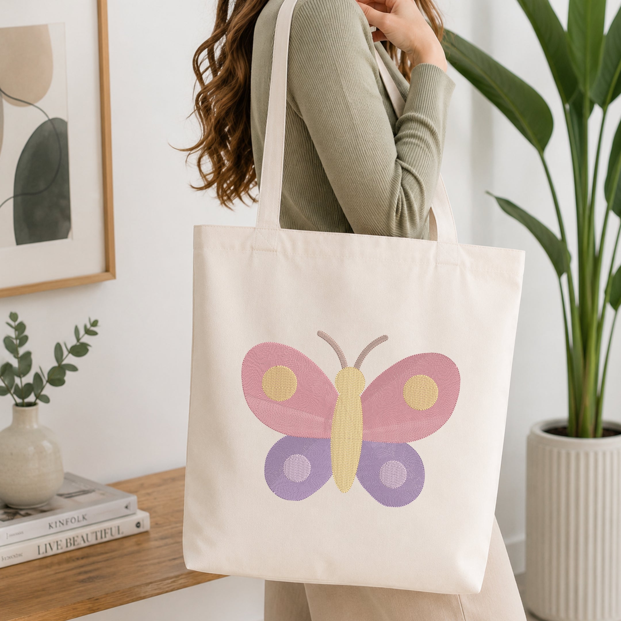 A woman wearing a sage green ribbed sweater carries a cream canvas tote bag with a colorful embroidered butterfly design in pink, purple, and yellow. The scene is set in a bright, modern room with a wooden table, books, a small plant, and a potted green plant in the background.