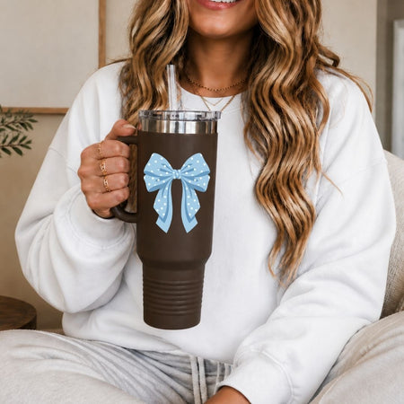 A woman in a white sweatshirt holds a brown insulated tumbler with a handle, clear lid, straw, and a light blue bow graphic on the front.