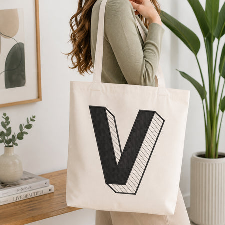 A woman wearing a sage green ribbed long-sleeve top carries a cream canvas tote bag with a large black 3D-style letter 'V' printed on the front, set in a bright, modern room with a wooden table, framed art, and a potted plant.