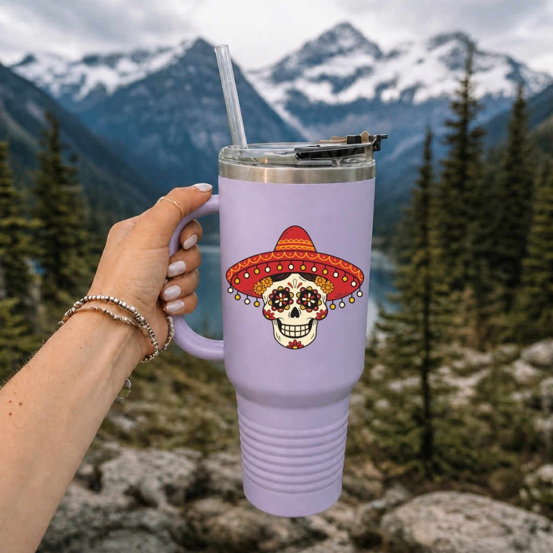 A hand holds a lavender insulated tumbler with a clear straw in front of a mountain and forest background. The tumbler features a sugar skull graphic wearing a red and orange sombrero.